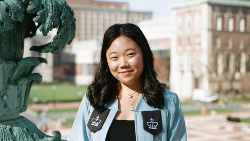 Young Asian woman, smiling with her mouth closed, dressed in Columbia University graduation gown. The background is the University
