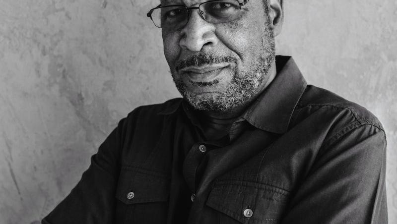 Older Black man with wire framed glasses, a scruffy beard, and his arms crossed across his chest