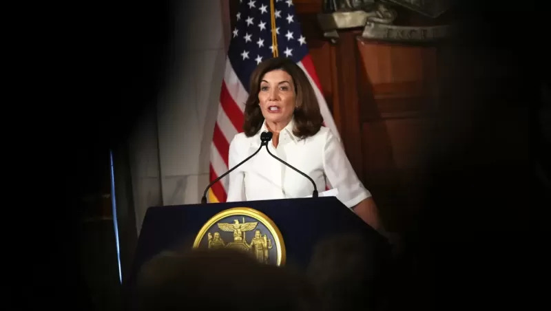 A woman in a white blouse with dark, should-length hair stands at a podium in front of an American flag.