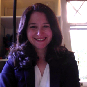 A woman smiling. She has shoulder-length hair. She is in front of a window.